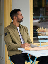 Model wearing FRAHM Lightweight Blouson Jacket 2026 Olive Green sat at outdoor table